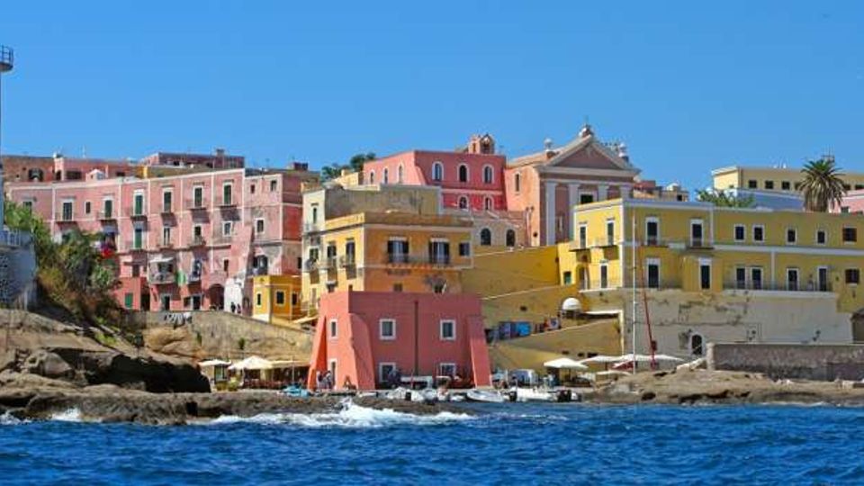 Vista costiera di un borgo sul mare con edifici color pastello su una scogliera rocciosa e un faro bianco sulla sinistra. In primo piano si vede l’acqua con piccole barche e un molo ai piedi delle case, sotto un cielo sereno.