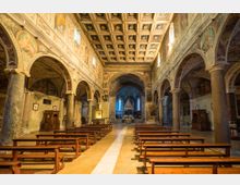 Interno di una chiesa storica con navata centrale fiancheggiata da colonne e archi, pareti affrescate e soffitto a cassettoni decorato. File di panche in legno conducono verso l’altare sul fondo, illuminato da finestre laterali.