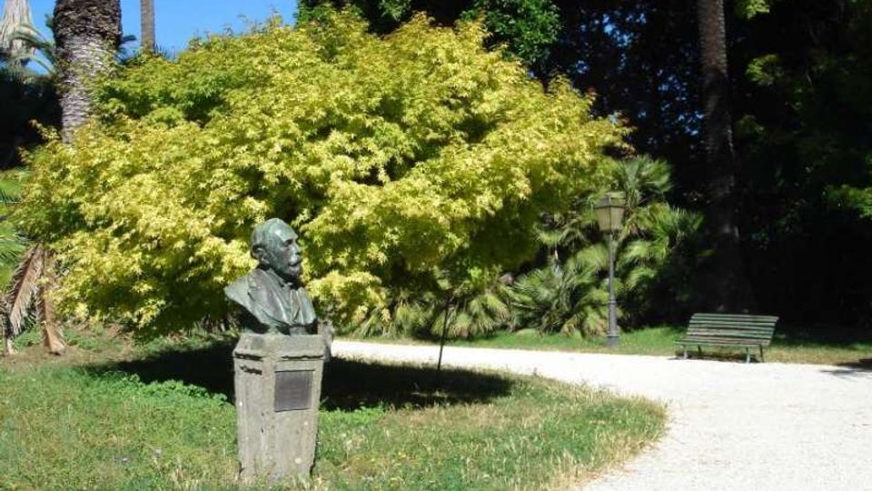 Parco con sentiero in ghiaia curvo tra prato e alberi, tra cui palme e un grande albero dalla chioma giallo-verde. In primo piano c’è un busto in bronzo su piedistallo in pietra, con panchina e lampione sullo sfondo.