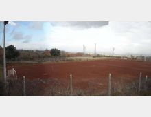 Campo da calcio in terra rossa recintato, con porte bianche e linee del terreno visibili. Sullo sfondo si vedono alberi, tralicci e un paesaggio rurale sotto un cielo nuvoloso.