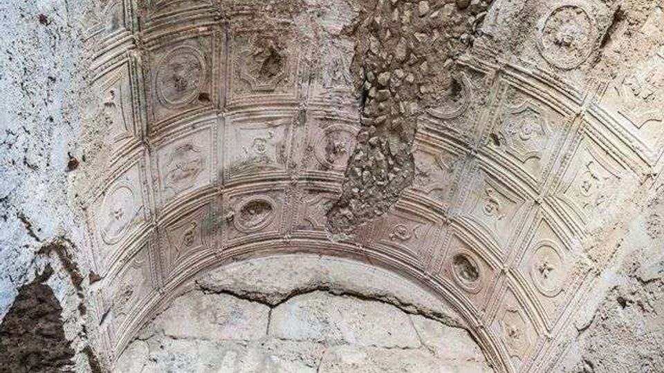 Soffitto a volta in pietra di un ambiente antico, con pannelli decorativi a cassettoni e rosoni scolpiti. Parte della volta è deteriorata, con intonaco mancante e pietrame a vista sopra un arco in muratura.