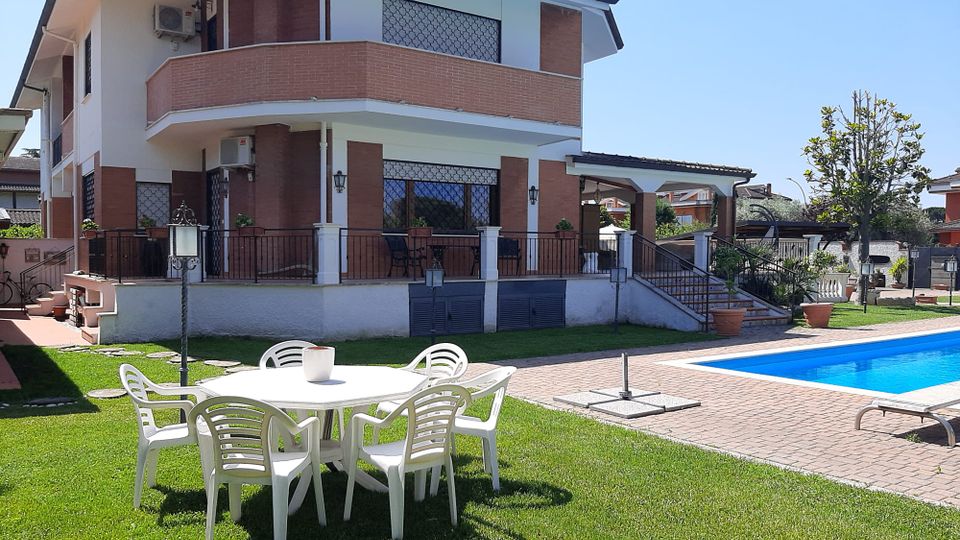 Villa a due piani con facciata in mattoni e intonaco bianco, terrazzi con ringhiere metalliche e finestre con grate decorative. In primo piano giardino con tavolo e sedie bianche su prato, vialetto in mattoni e piscina rettangolare sul lato destro.