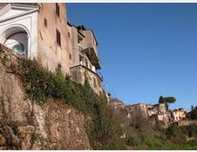 Case in pietra e intonaco costruite sul bordo di una scarpata rocciosa, con muri di contenimento e vegetazione spontanea sul pendio. Sullo sfondo si vede un piccolo borgo collinare con altri edifici, un grande albero e cielo sereno.
