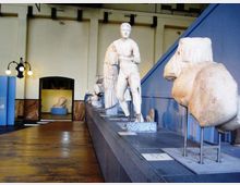 Sala museale con pavimento in legno e pareti chiare, con una lunga pedana espositiva che ospita statue e frammenti scultorei in marmo. In primo piano si vedono un torso e una statua classica, mentre sullo sfondo sono presenti un lampione a più luci e un’apertura verso un’altra sala.