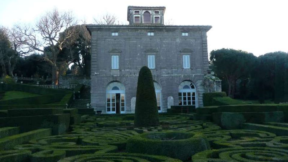 Facciata di una villa in pietra a due piani con grandi porte ad arco e finestre simmetriche, con un piccolo belvedere sul tetto. In primo piano si vede un giardino formale con siepi potate a disegni geometrici e un alto cespuglio centrale, circondato da alberi e prati.
