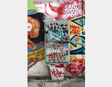 Armadio tecnico metallico da strada ricoperto di graffiti e tag, posizionato davanti a un muro con murales multicolori e scritte spray. In primo piano si vede il marciapiede in cemento e mattonelle con piccoli detriti.