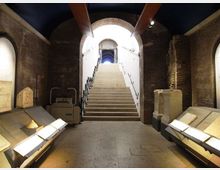 Interno di uno spazio museale in mattoni con soffitto a volta e ampio arco centrale, dominato da una scalinata che conduce a un’uscita illuminata. Ai lati della sala ci sono teche e basamenti con reperti esposti lungo le pareti.