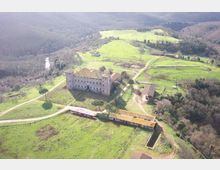Veduta aerea di un grande edificio storico in pietra con torre, circondato da prati verdi e strade sterrate in una campagna collinare. Intorno si vedono campi, piccoli edifici rurali e un’area boschiva ai margini.