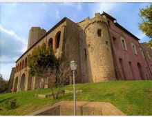 Edificio storico in pietra simile a un castello, con torre cilindrica e una lunga facciata ad archi, affiancato da una sezione intonacata rosa con finestre. In primo piano c’è un pendio erboso con alberi e un lampione, sotto un cielo parzialmente nuvoloso.