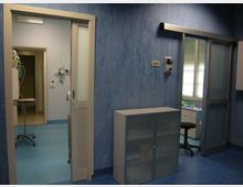 Interno di una struttura sanitaria con pavimento blu e pareti azzurre, con porte scorrevoli e un armadietto metallico. Sullo sfondo si vedono un corridoio con attrezzature mediche e una piccola stanza con finestra e arredi da lavoro.