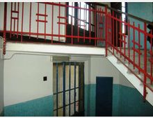 Interno di un edificio con scala e pianerottolo, caratterizzato da una ringhiera metallica rossa geometrica e pareti rivestite a metà con piastrelle turchesi. In basso si vede un corridoio con una porta chiusa e un’apertura con inferriata metallica, illuminata da finestre alte sul fondo.