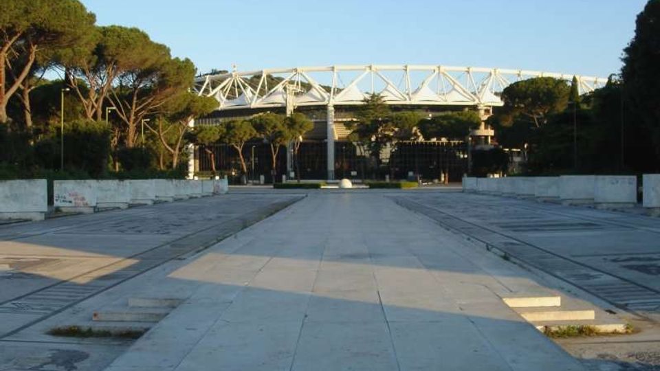 Ampio piazzale pavimentato in cemento con gradoni bassi e aiuole laterali, circondato da alberi alti. Sullo sfondo si vede uno stadio con struttura metallica bianca sopra le tribune, dietro una recinzione.