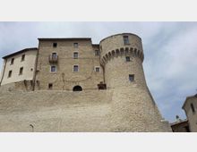 Castello in pietra con alta cinta muraria e torre cilindrica merlata, affacciato su un cielo nuvoloso. La struttura presenta facciate in muratura e intonaco con piccole finestre e balconcini, visibile da una prospettiva dal basso.