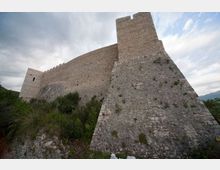Mura esterne di un castello o fortezza in pietra con torre e bastioni inclinati, viste dal basso lungo un pendio roccioso. Vegetazione bassa intorno alla base e cielo nuvoloso sullo sfondo.