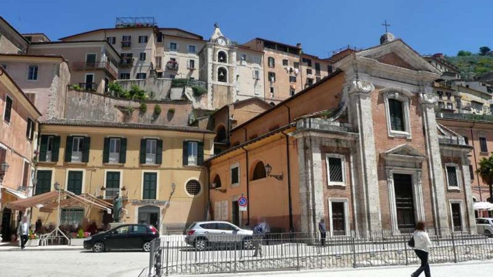 Piazza urbana con una chiesa in stile classico in primo piano, con facciata in mattoni e pietra, portale centrale e timpano sormontato da una croce. Sullo sfondo si vedono edifici residenziali dai colori chiari disposti su un pendio, con un campanile e cielo azzurro sopra.
