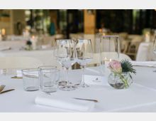 Tavolo apparecchiato in un ristorante elegante con tovaglia bianca, bicchieri da vino e da acqua, posate dorate e un piccolo centrotavola floreale in vaso di vetro. Sullo sfondo si intravedono altri tavoli e grandi vetrate in un ambiente luminoso.