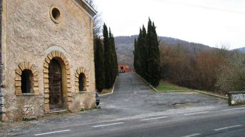 Piccola cappella in pietra con portale e finestre ad arco e oculo sulla facciata, affacciata su una strada asfaltata. Accanto parte un viale in lieve salita fiancheggiato da cipressi che conduce verso edifici e colline boscose sullo sfondo.