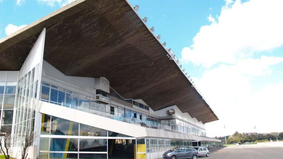 Edificio moderno a più livelli con ampie vetrate e una grande copertura sporgente inclinata, affacciato su un’ampia area asfaltata tipo parcheggio. In primo piano si vedono una striscia di prato e alcune auto, con cielo azzurro sullo sfondo.