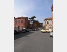 Ampio parcheggio asfaltato tra edifici bassi in stile tradizionale con facciate colorate e finestre con persiane. Sullo sfondo si vedono altri fabbricati e un grande albero, con auto parcheggiate lungo i lati sotto un cielo sereno.