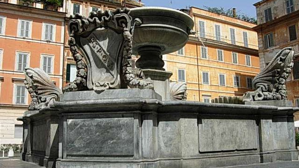 Fontana monumentale in pietra con grande vasca circolare centrale, base rettangolare e sculture decorative ai lati, collocata in una piazza urbana. Sullo sfondo si vedono palazzi storici a più piani con facciate colorate e finestre regolari.