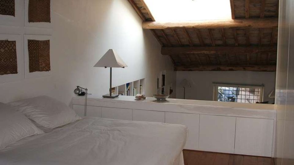 Camera da letto in soppalco con letto matrimoniale, armadiature bianche basse e lampada da tavolo. Soffitto mansardato con travi in legno e grande lucernario, pavimento in legno e finestra sulla parete di fondo.