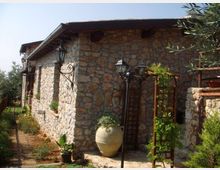 Edificio rustico in pietra con tetto sporgente e lanterne in ferro battuto fissate alla parete, lungo un vialetto in mattoni. All’esterno si vedono un grande vaso in terracotta, una struttura a graticcio con piante rampicanti e vegetazione da giardino ai lati.
