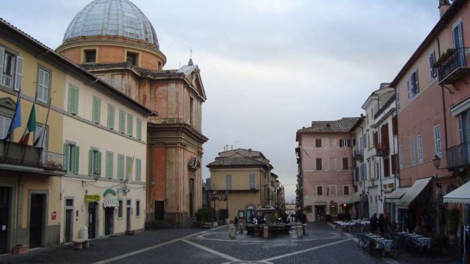 Piazza lastricata in pietra con fontana centrale, circondata da edifici storici a più piani con persiane e balconi. Sul lato sinistro domina una grande chiesa con facciata in pietra e ampia cupola.