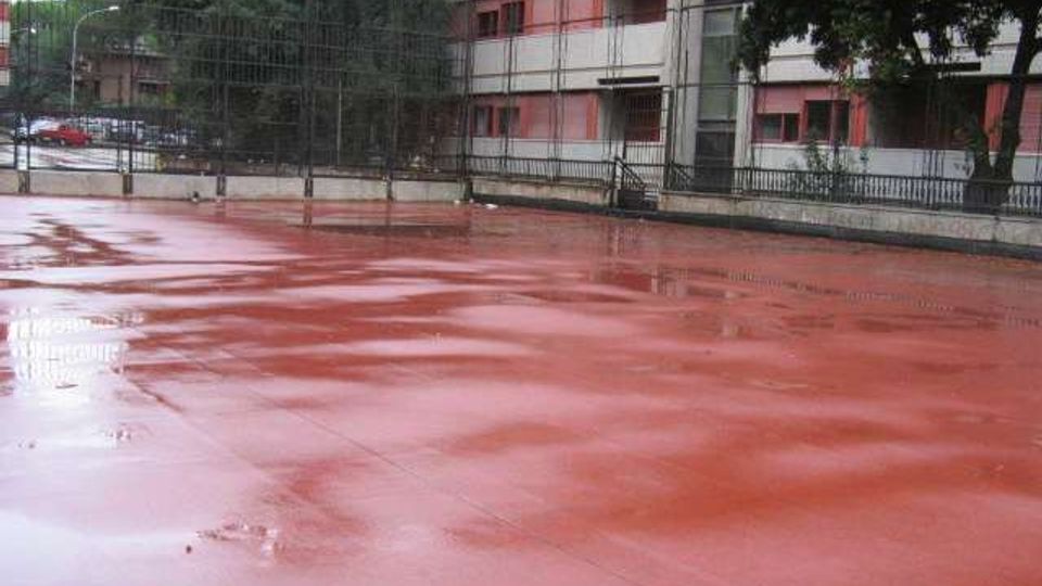 Campo sportivo all’aperto con pavimentazione rossa bagnata dalla pioggia e recinzione metallica alta lungo il perimetro. Sullo sfondo si vedono alberi e un edificio residenziale con balconi e finestre.