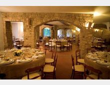 Sala da pranzo in stile rustico con grandi archi e pareti in pietra a vista, pavimento in terracotta e illuminazione calda. Tavoli rotondi apparecchiati con tovaglie e sedie in legno disposti in più aree sotto le arcate.