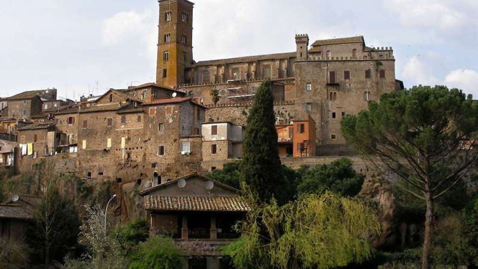 Borgo storico su un’altura con edifici in pietra e una grande chiesa o complesso fortificato con alta torre campanaria. In primo piano si vede un’area verde con alberi e un piccolo edificio con tetto in coppi, sullo sfondo cielo parzialmente nuvoloso.