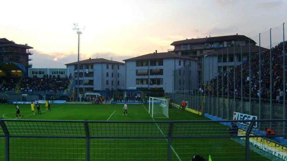 Campo da calcio di uno stadio di quartiere con tribune laterali recintate e illuminazione a torre; sullo sfondo si vedono palazzine residenziali e un cielo al tramonto. In primo piano sono visibili la rete metallica di separazione e un’area vicino alla porta con bandierina d’angolo.