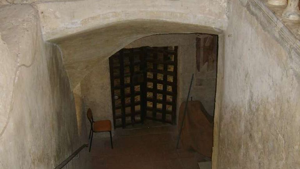 Scala interna in pietra che scende verso un ambiente sotterraneo con soffitto ad arco; in alto si vede una balaustra con colonnine. In fondo c’è una pesante porta a grata in legno, con pareti in muratura grezza e illuminazione soffusa.