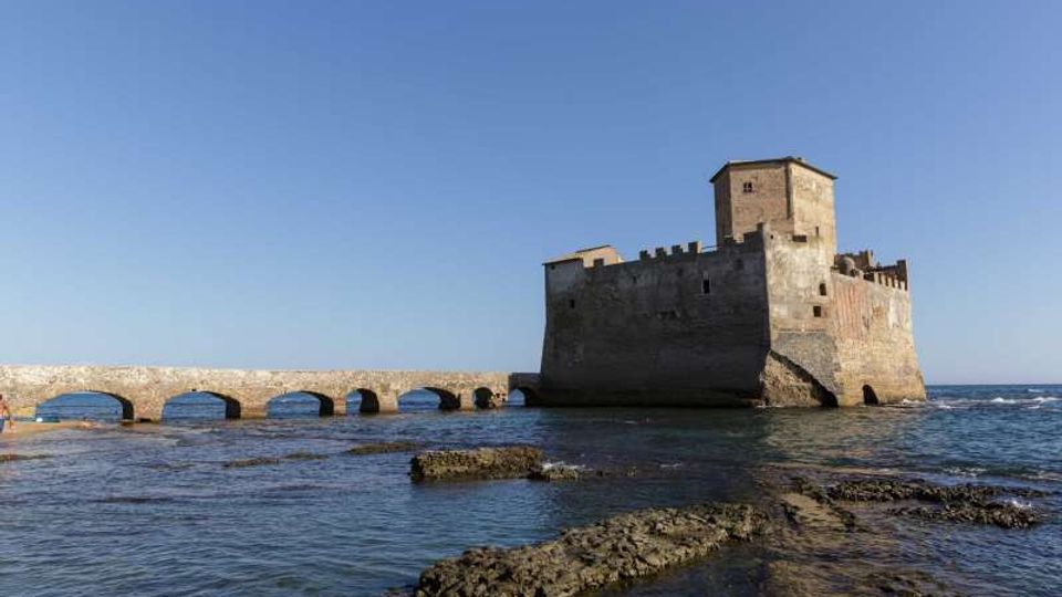 Fortezza costiera in pietra su un isolotto, con torre quadrata e mura merlate, collegata alla riva da un ponte basso ad archi. In primo piano si vede il mare calmo con scogli affioranti e un ampio cielo sereno sullo sfondo.