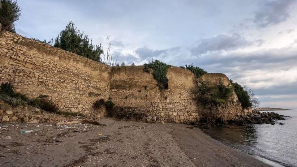 Antiche mura in pietra lungo una spiaggia sabbiosa e ghiaiosa, con vegetazione in cima e tratti erosi vicino alla riva. Il mare calmo si estende sulla destra sotto un cielo nuvoloso.