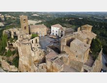 Veduta aerea di un borgo medievale in pietra con rovine di mura e torri, una piazza centrale e edifici restaurati con tetti in coppi. Intorno si estendono boschi e campi collinari, con una strada che costeggia il complesso sul lato destro.