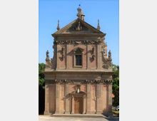 Facciata frontale di una chiesa in mattoni con elementi decorativi in pietra, portale centrale e finestra sopra l’ingresso. Il prospetto è sormontato da un timpano con statue e pinnacoli, su sfondo di cielo sereno e alberi ai lati.