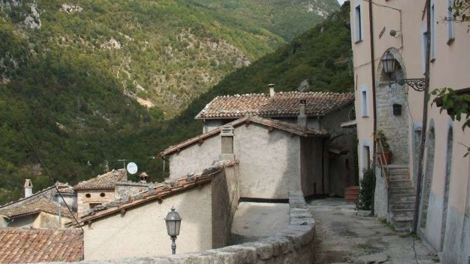 Vicolo acciottolato in un borgo di montagna, con case in pietra intonacata e tetti in coppi, muretto curvo e lampione. Sullo sfondo si vedono colline boscose e montagne sotto un cielo nuvoloso.