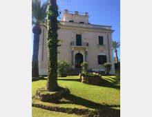 Facciata di una villa storica a tre livelli color beige con ingresso ad arco, balcone centrale e persiane verdi. Davanti si estende un prato curato con palme e aiuole in pietra, sotto un cielo sereno.