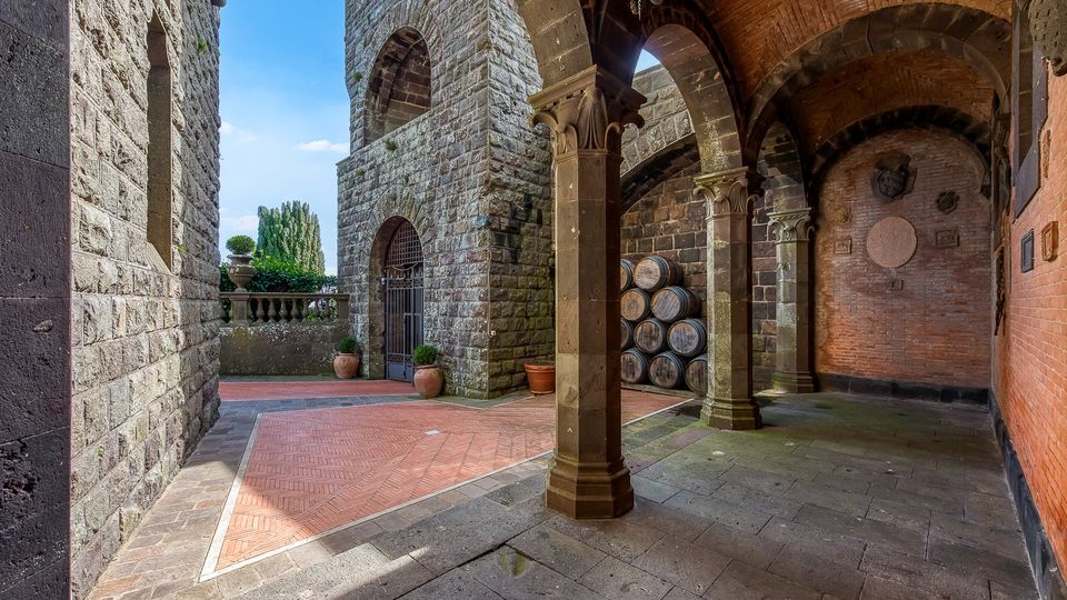 Cortile in pietra di un edificio storico con portico ad archi sostenuto da colonne e soffitto a volte in mattoni. Sul lato destro ci sono botti accatastate e una parete in mattoni, mentre a sinistra si vedono mura in pietra, un cancello ad arco e una balaustra con vasi.