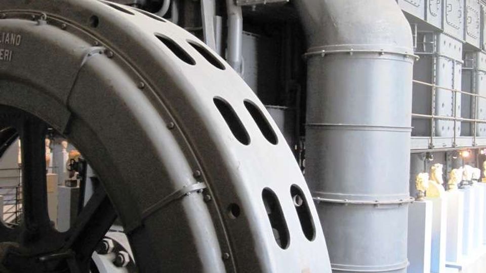 Grande sala industriale con un enorme macchinario metallico grigio, con carter curvo forato, tubazioni e passerella superiore con ringhiera. Sullo sfondo si vedono pannelli e condotti di grandi dimensioni allineati lungo la parete, con pavimento in piastrelle e griglie di aerazione.