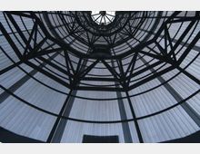 Vista dal basso di una cupola circolare in vetro e metallo, con travi nere che formano una struttura reticolare radiale. Il soffitto traslucido crea un lucernario centrale e anelli concentrici che definiscono l’architettura interna.