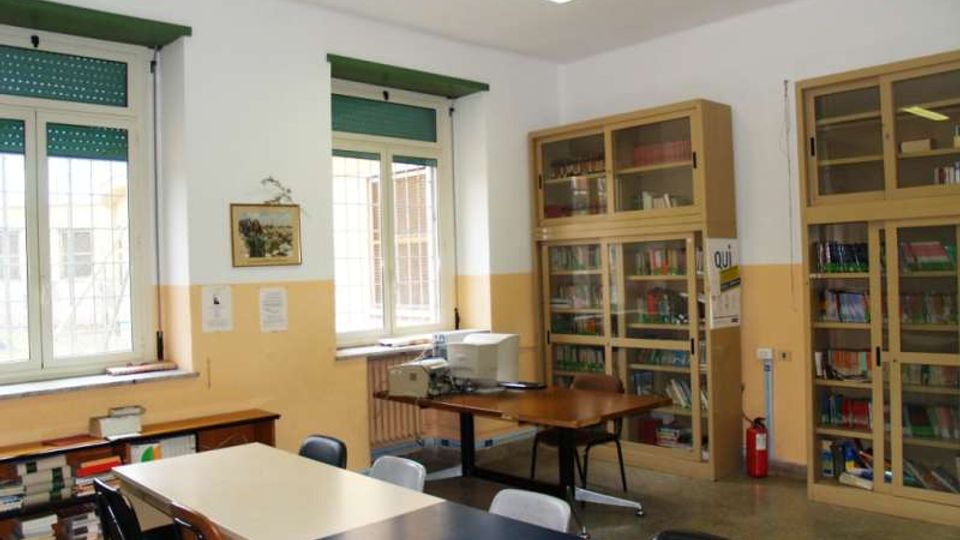 Aula scolastica con tavoli e sedie, pareti bianche con fascia gialla e due grandi finestre con tapparelle verdi. Sul lato destro ci sono armadi vetrati pieni di libri e materiali, con una stampante su un tavolo e un estintore vicino alla parete.
