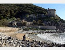 Piccola spiaggia con ciottoli e riva sabbiosa affacciata sul mare, con onde che arrivano a riva e un camminamento costiero protetto da muretti in pietra. Sul pendio roccioso e boscoso retrostante si vede un complesso fortificato con una torre circolare in pietra in cima.