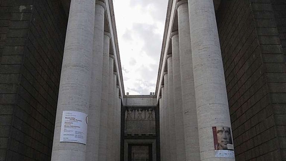 Facciata monumentale di un edificio pubblico con due alte file simmetriche di colonne cilindriche in pietra chiara, affiancate da pareti scure e raggiunte da un’ampia scalinata. Al centro si vede un ingresso incassato con porta vetrata, sotto un cielo nuvoloso.