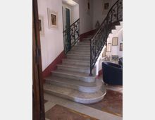 Scala interna in marmo con ampi gradini curvi e ringhiere in ferro battuto decorato, all’interno di un edificio con pareti bianche e fascia bordeaux. Sulle pareti sono appesi quadri e cornici, con una piccola area salotto e pavimento in marmo policromo.