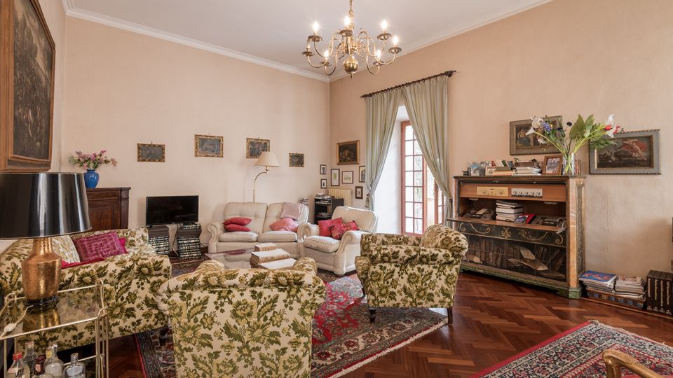 Soggiorno in stile classico con parquet a spina di pesce, tappeti persiani e lampadario centrale, arredato con divani chiari e poltrone floreali attorno a un tavolino. Una portafinestra con tende chiare illumina la stanza; alle pareti sono appesi quadri e sul lato destro c’è un mobile con libri e oggetti decorativi.