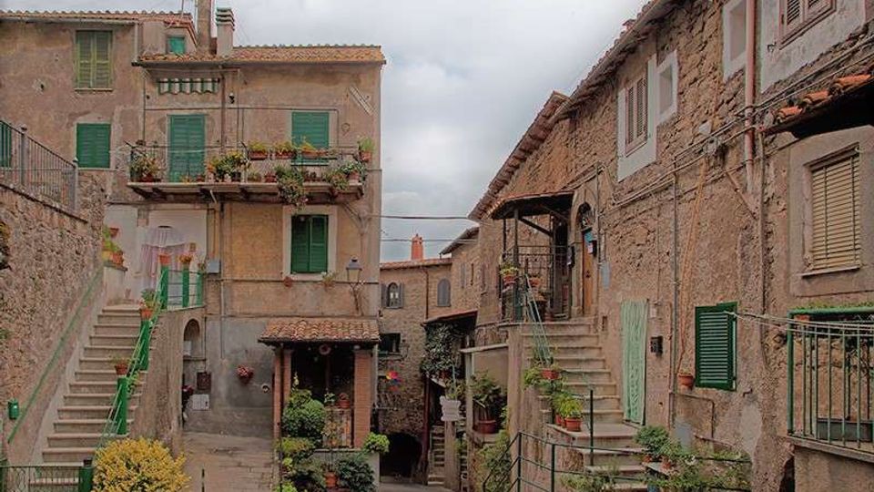 Cortile in un borgo con case in pietra e intonaco, persiane verdi, balconi con fioriere e scale esterne con ringhiere. Pavimentazione in pietra, vasi di piante e piccoli ingressi sotto portici, con cielo nuvoloso sullo sfondo.