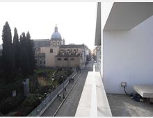 Vista urbana da una terrazza con parapetto bianco su una strada pedonale sottostante; sullo sfondo si vede un edificio storico con cupola e facciate ad archi. A sinistra c’è un’area verde recintata con alti alberi, mentre a destra si notano pareti e sporgenze di un edificio moderno.