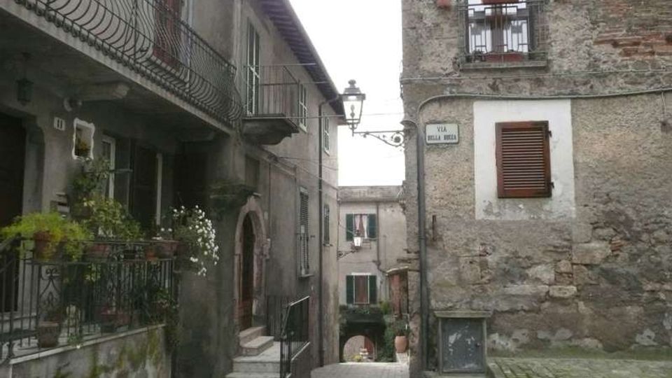 Vicolo acciottolato in un borgo storico, stretto tra edifici in pietra e intonaco con balconi in ferro battuto, persiane in legno e un lampione a muro. In fondo si vede un passaggio ad arco e una targa stradale su una facciata.