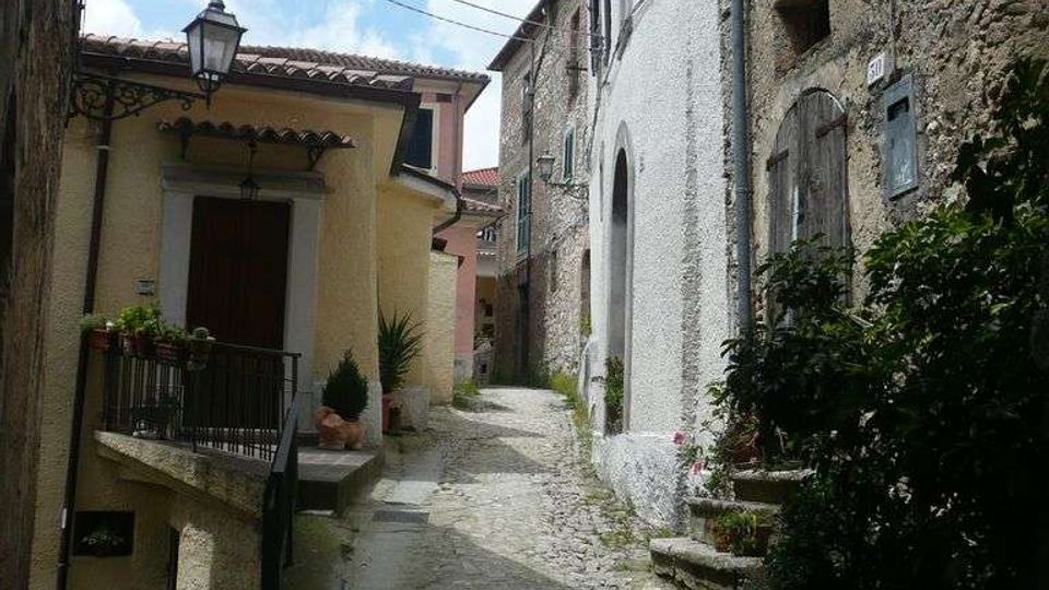 Vicolo stretto in pietra con pavimentazione acciottolata che sale tra case in muratura e intonaco, con finestre ad arco e piccoli balconi. Lampione a parete, cavi sospesi e vasi di piante lungo i muri sotto un cielo parzialmente nuvoloso.